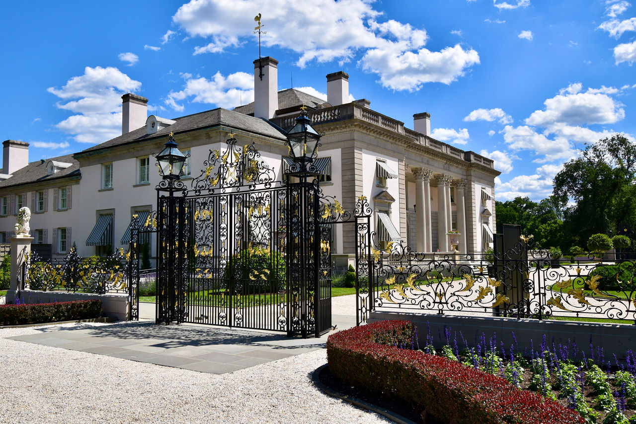 Outside the estate on a sunny day, the tall iron gates at the main entrance with the Mansion building behind it and blooming gardens all around. 