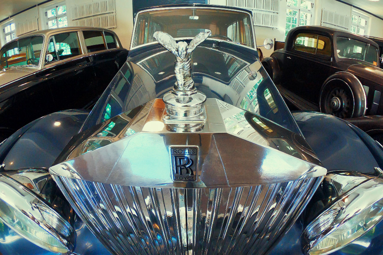 A close-up of a Rolls-Royce hood ornament with a custom "Blueberry Blue" color is parked between two classic cars. 