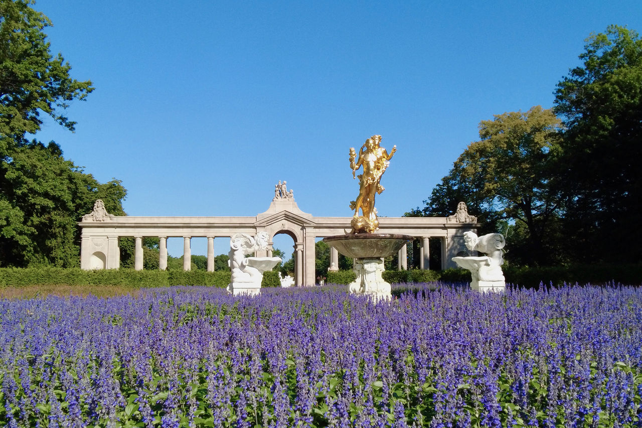 Purple flowers bloom all around a gold leaf statue with a stone building in the background.