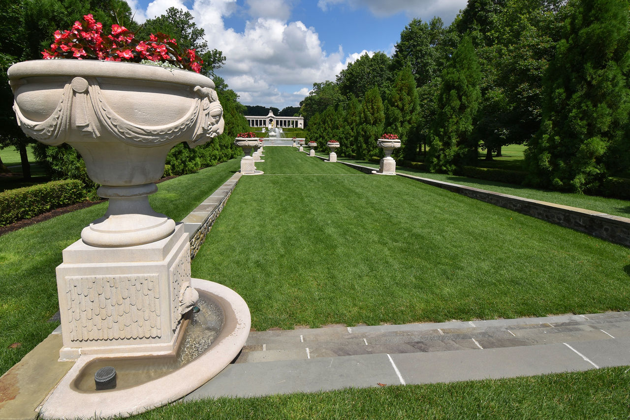 The Long Walk at Nemours Estate with a sunny stretch of green grass lined by trees and rectangled terraced lawns, and a large white planter with red blooms in the foreground.  