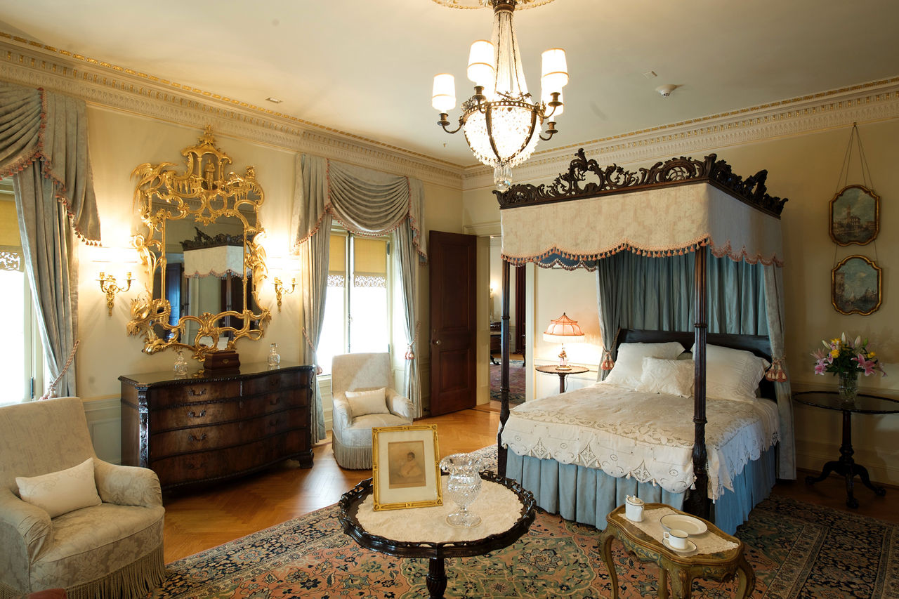 The duPont bedroom with a four-poster bed, gold mirror, chandelier, and many layers of window and bed fabrics in white and gold. 