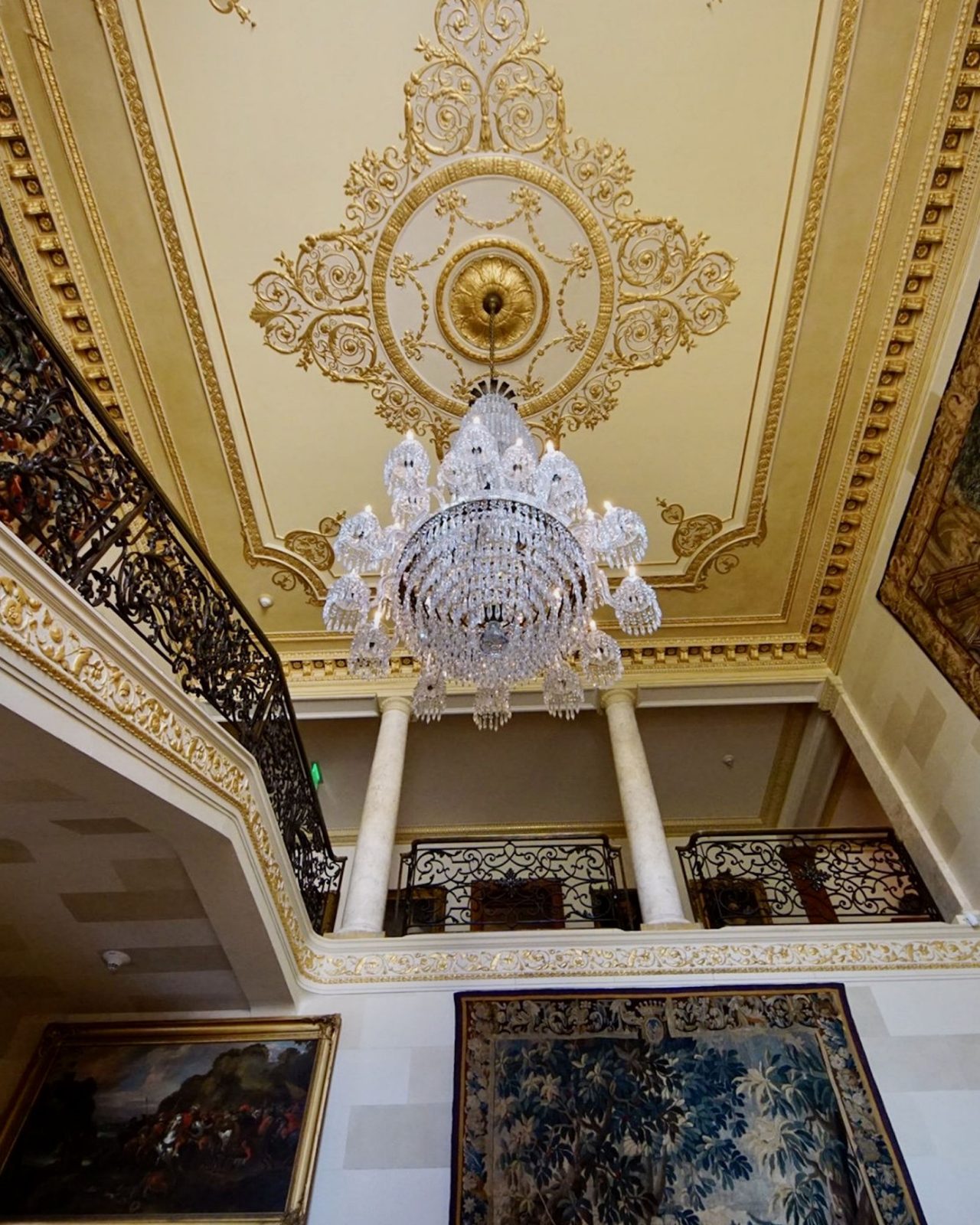 A "featured" chandelier in Stair Hall is hanging next to an iron bannister from a royal yellow ceiling. 