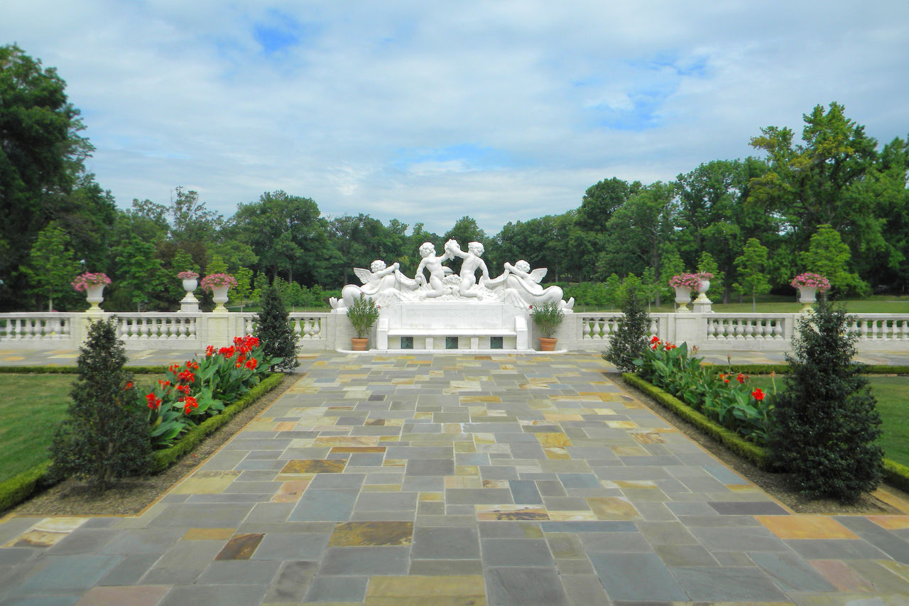 A sunny afternoon outside in the Gardens on the stone walkway with flowers blooming and a statue with a bench at the end.